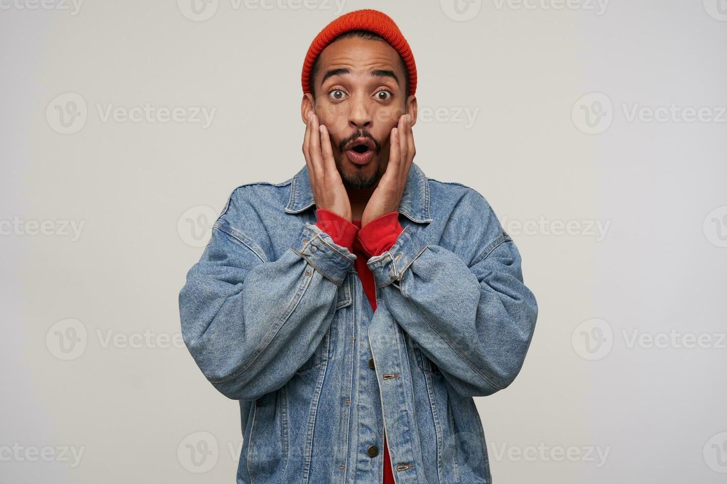Shocked young handsome dark skinned brunette guy with beard holding face with palms and rounding eyes with raised eyebrows while standing over white background photo