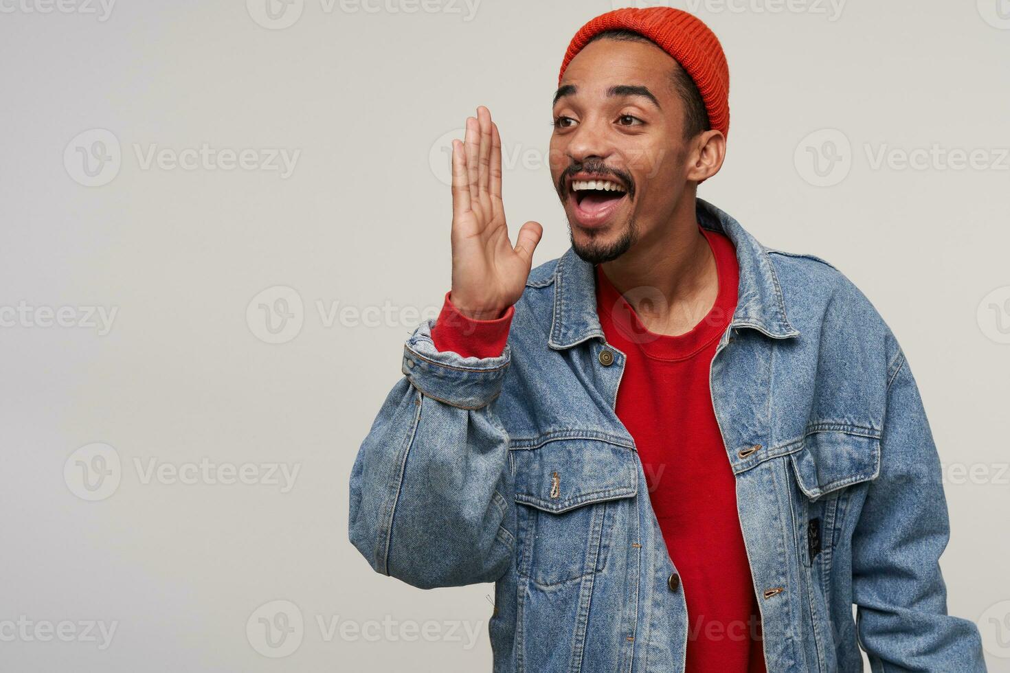 Indoor photo of cheerful lovely young bearded dark skinned guy dressed in red hat, red pullover and jeans smiling widely with raised palm while posing over white background