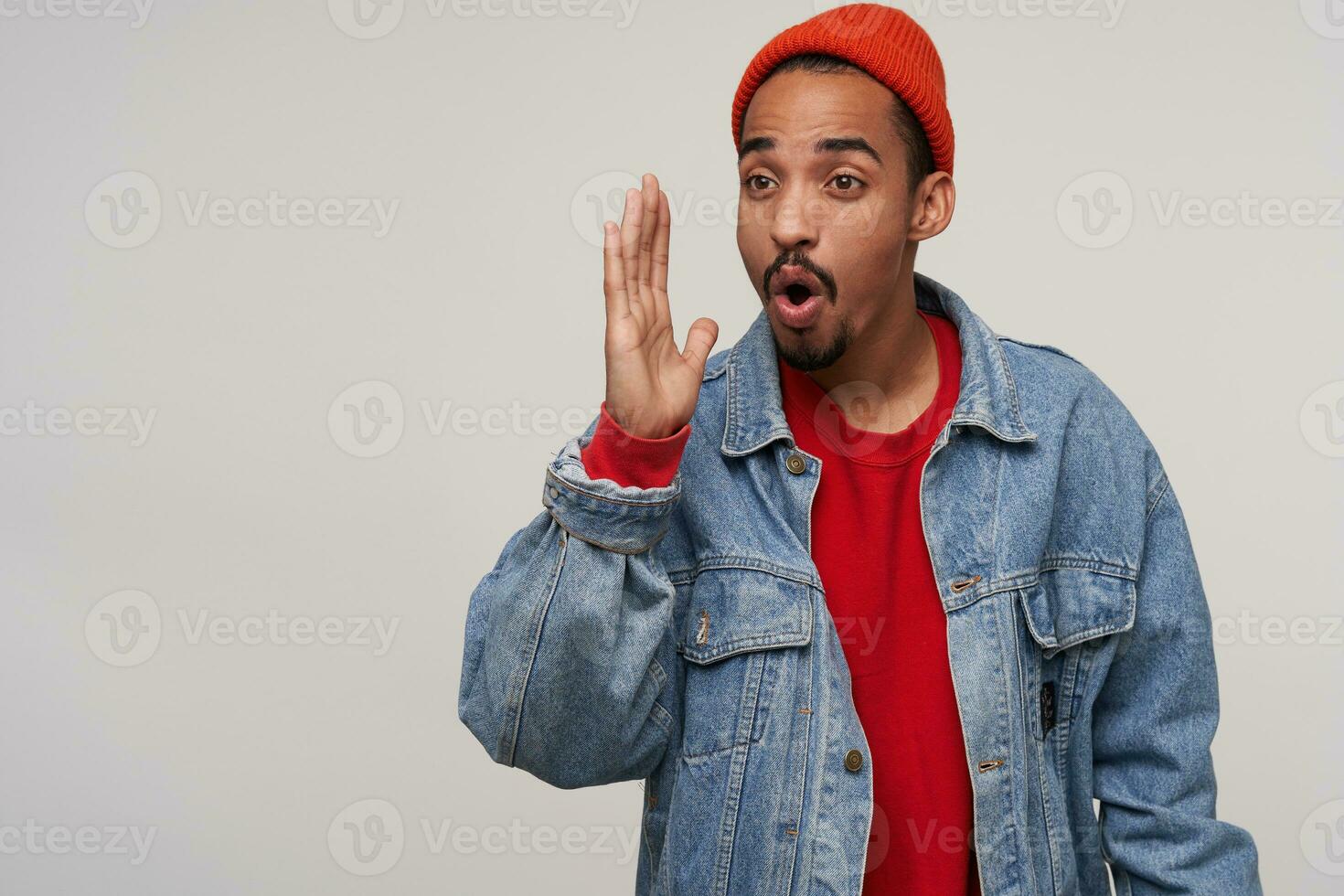 Portrait of young dark haired bearded man with dark skin rounding his mouth while looking ahead and keeping hand raised, standing against white background in casual clothes photo