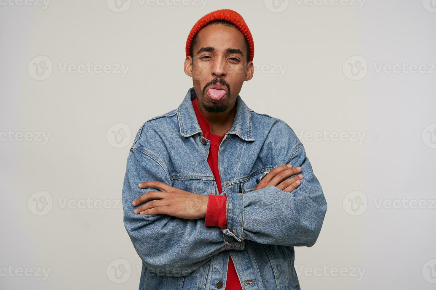 Indoor shot of offended young handsome bearded dark skinned guy squinting his brown eyes and showing tongue to camera, standing over white background with folded arms photo