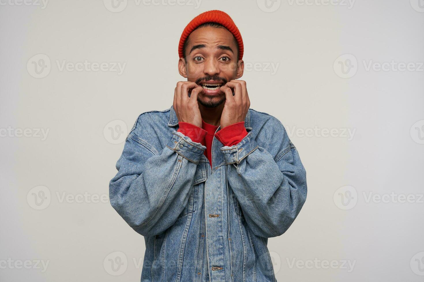 Frightened young pretty bearded dark skinned brunette male looking scaredly to camera and biting his hands, wrinkling forehead while posing over white background photo