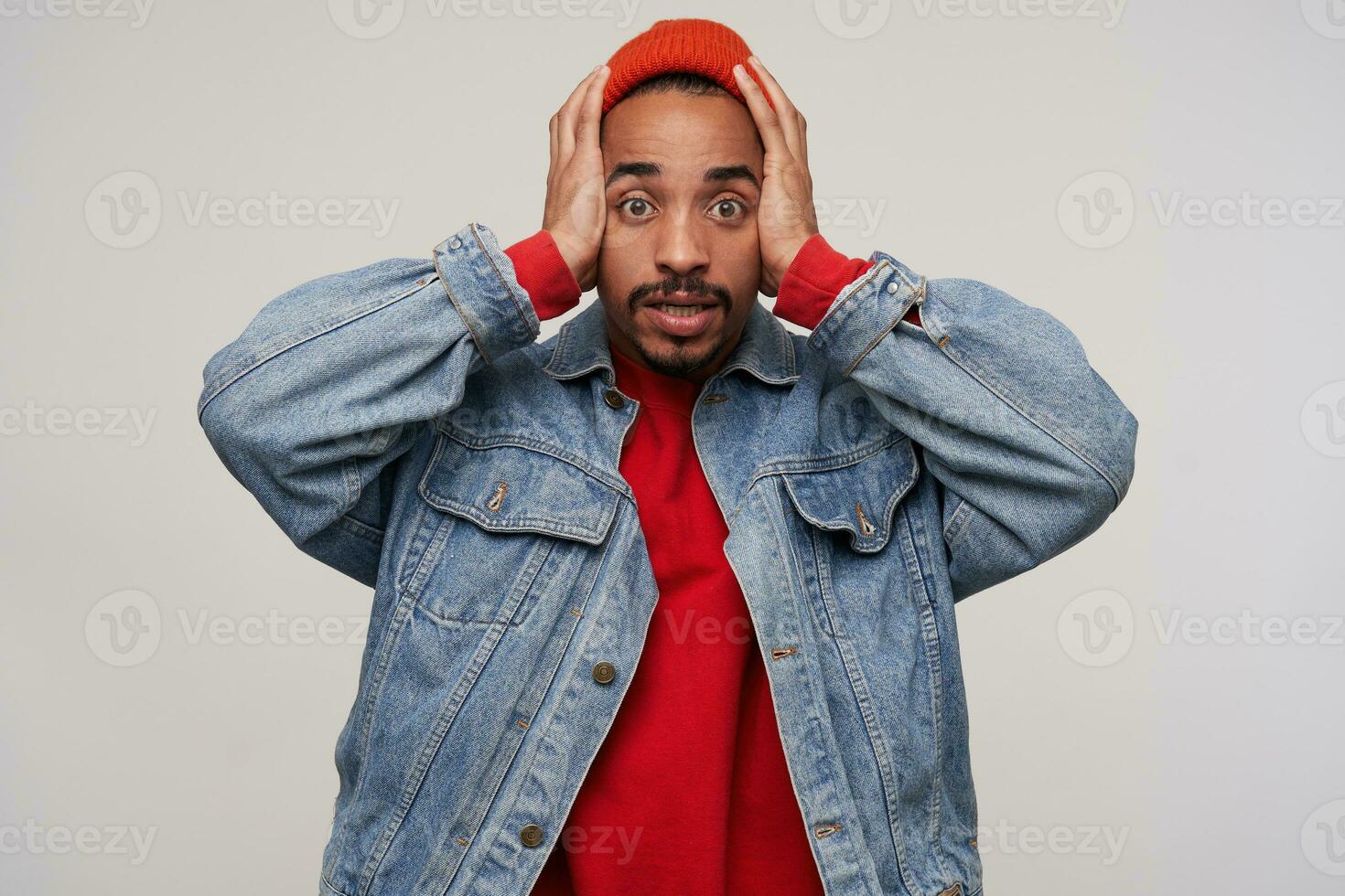 Bewildered young pretty dark haired bearded man with dark skin clutching his head with raised hands and looking perplexedly at camera, dressed in casual wear over white background photo