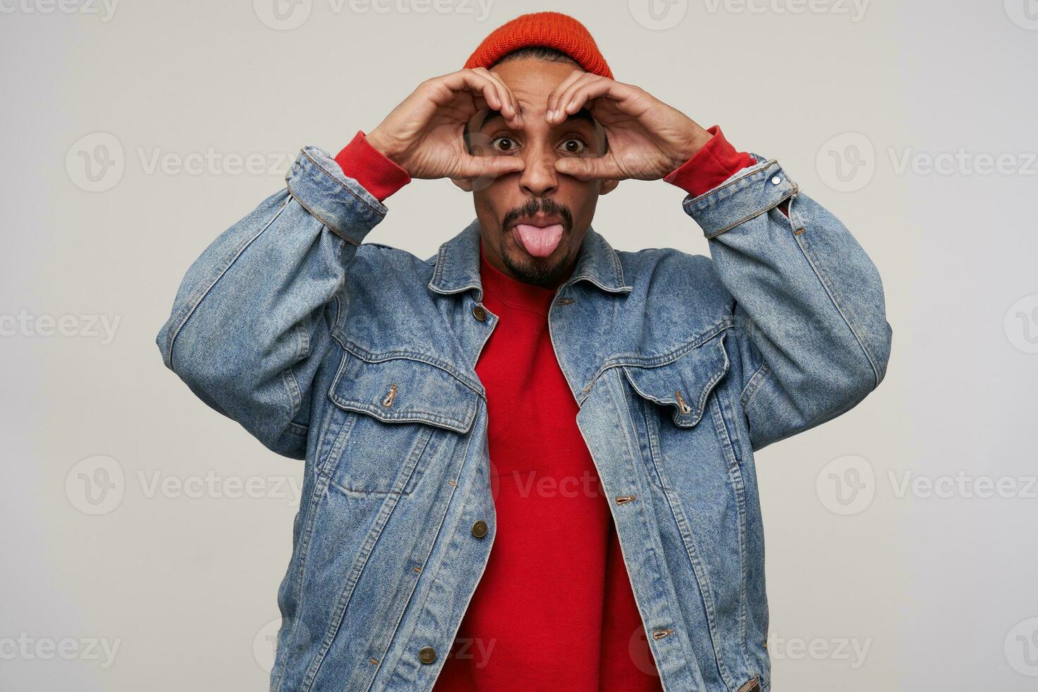 Funny shot of pretty young bearded dark skinned male showing tongue and making ridiculous faces while standing over white background in red hat, red pullover and jeans coat photo