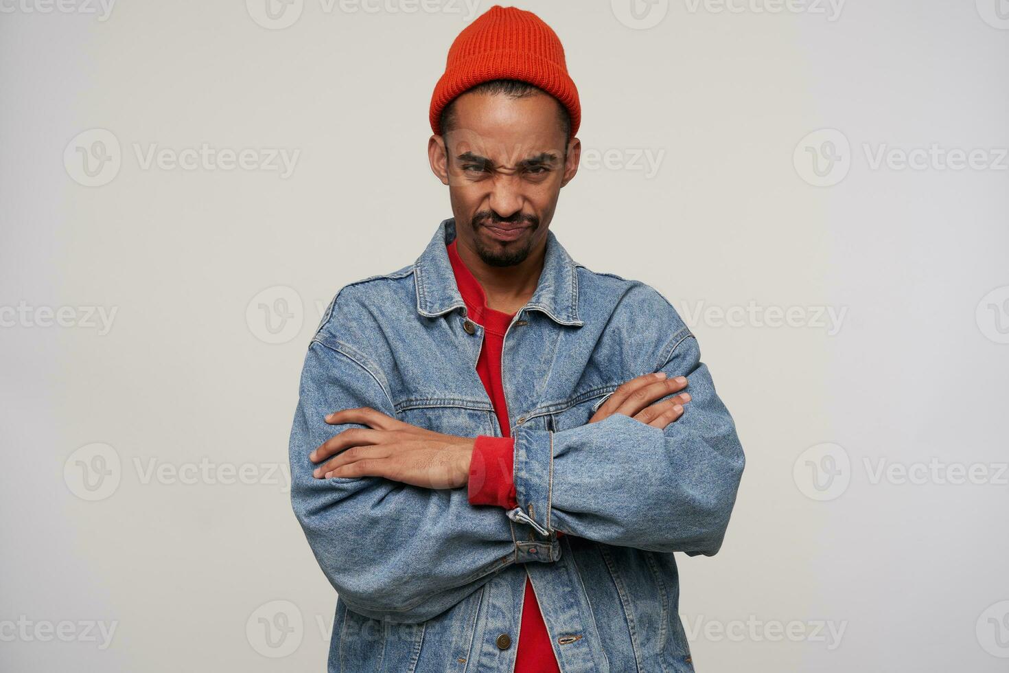 Displeased young dark haired bearded hipster with dark skin crossing hands on his chest and looking to camera with pout, frowning eyebrows with folded lips while posing over white background photo