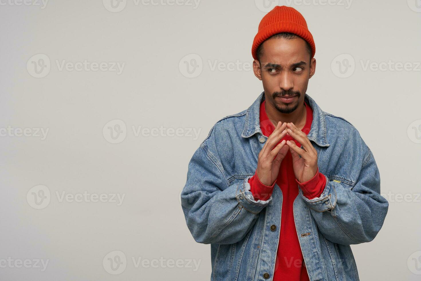 Pensive young lovely dark skinned bearded brunette man keeping hands together while plotting something with cunning face, dressed in casual wear while posing over white background photo