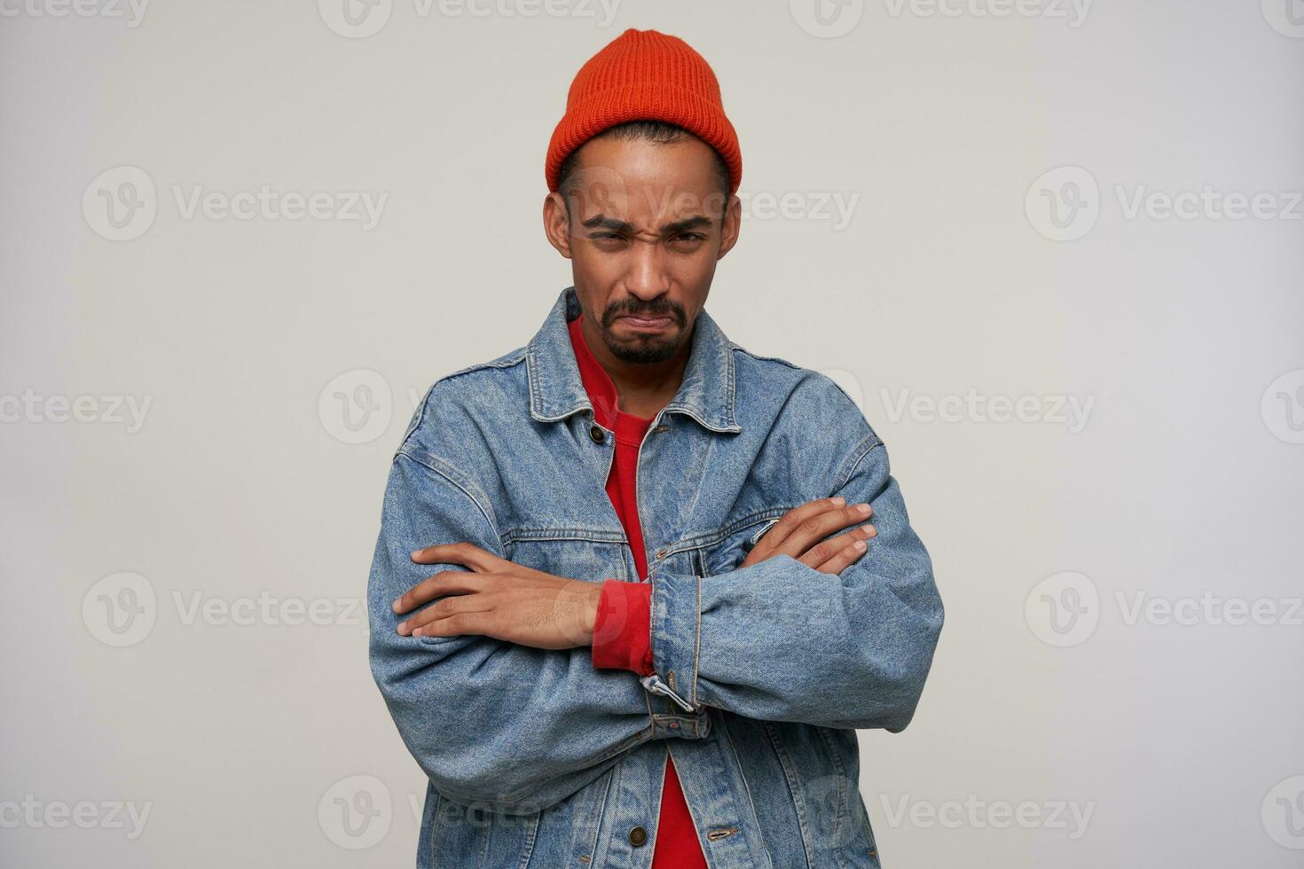 Offended young dark skinned brunette male folding hands on his chest while standing over white background, squinting and pursing lips while looking at camera photo