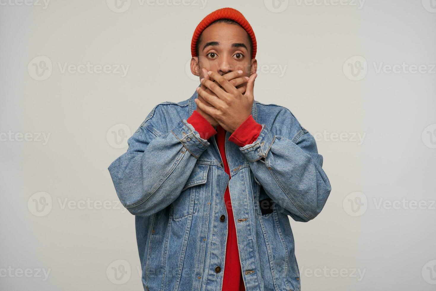 Photo of frightened young brown-eyed dark skinned bearded guy covering his mouth with raised palms while looking scaredly at camera, standing against white background