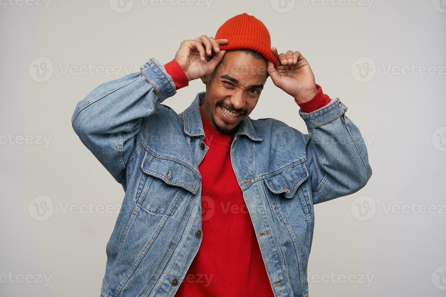 Cheerful young pretty dark skinned brunette male with beard holding his red cap with raised hands while standing over white background, being in nice mood and smiling happily photo