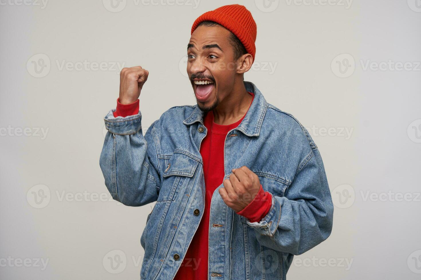 Overjoyed young dark skinned bearded brunette guy screaming happily with raised fists, wearing red cap, pullover and blue jeans coat while standing over white background photo