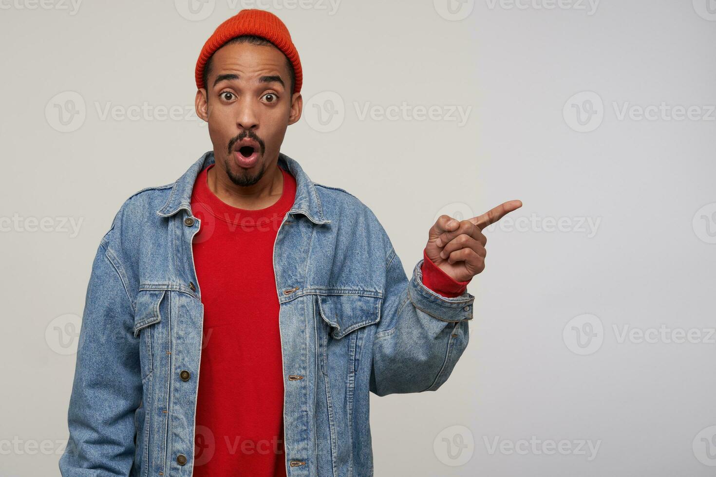Amazed young handsome dark skinned bearded brunette male pointing aside with wide eyes and mouth opened, raising surprisedly eyebrows while posing over white background photo