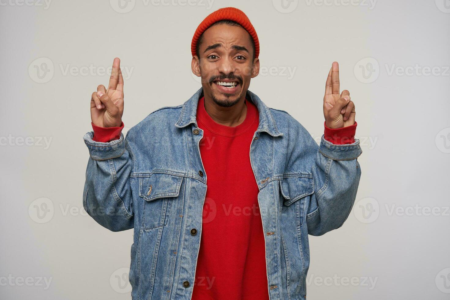 Hoping young brown-eyed dark skinned bearded male frowning his eyebrows while looking worringly at camera, raising hands with crossed fingers while posing over white background photo