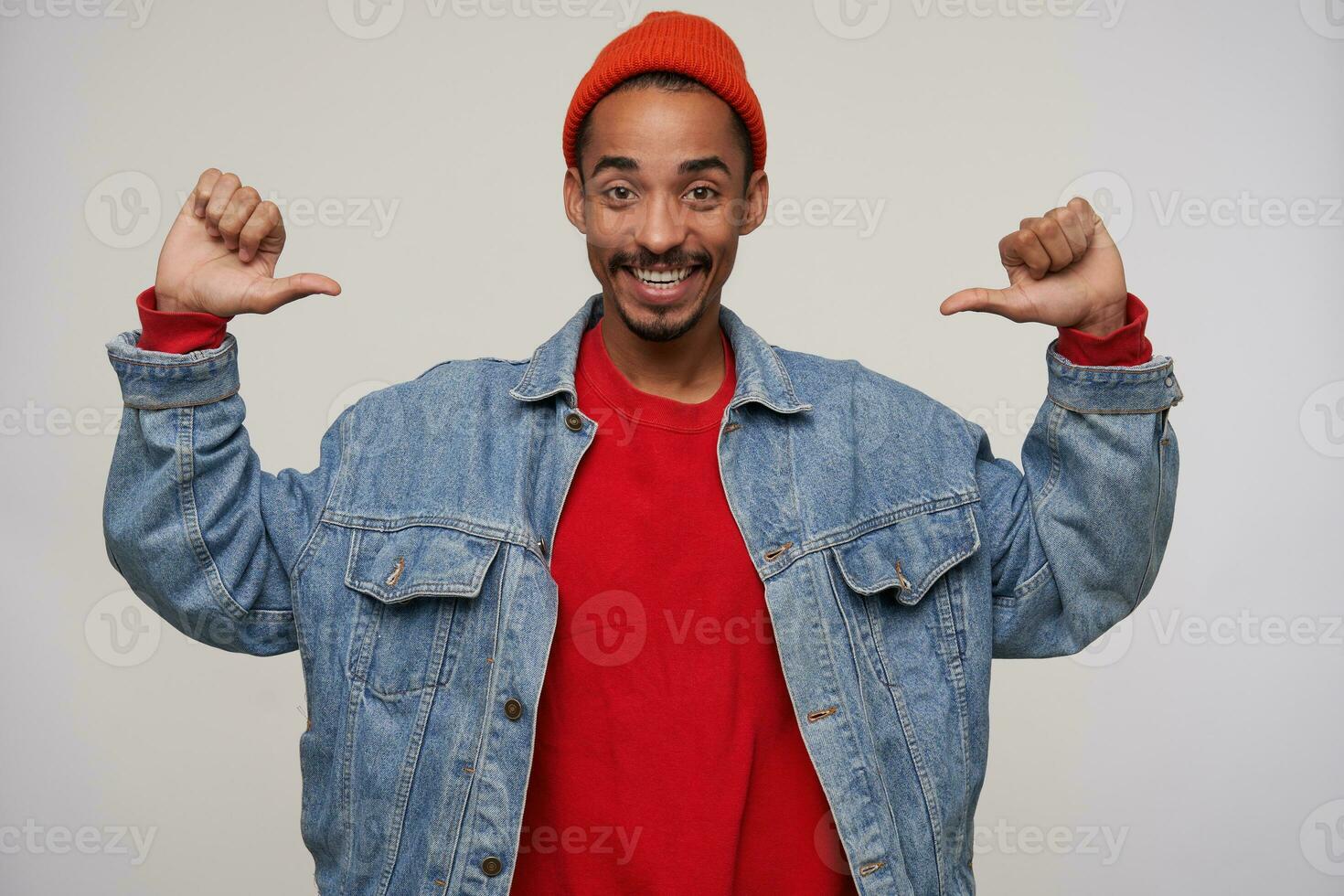 Happy young attractive dark skinned bearded brunette guy showing happily on himself with thumbs, smiling widely while at camera while posing over white background photo