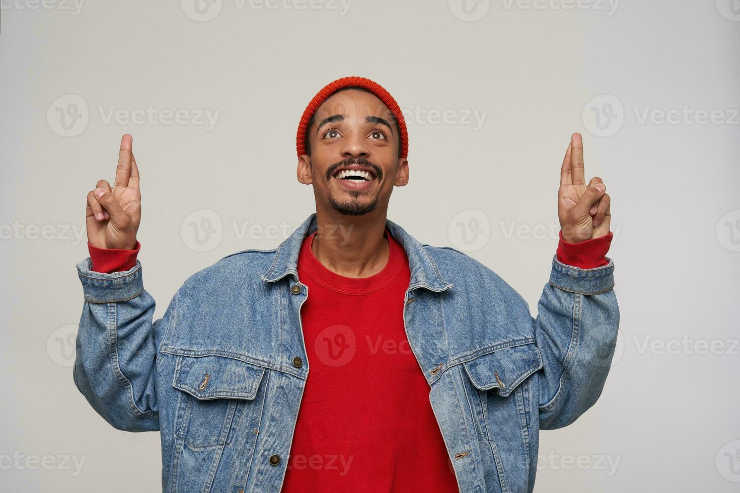 Happy young handsome bearded brunette male with dark skin looking hopefully upwards and keeping fingers crossed, dressed in casual clothes while posing over white background photo