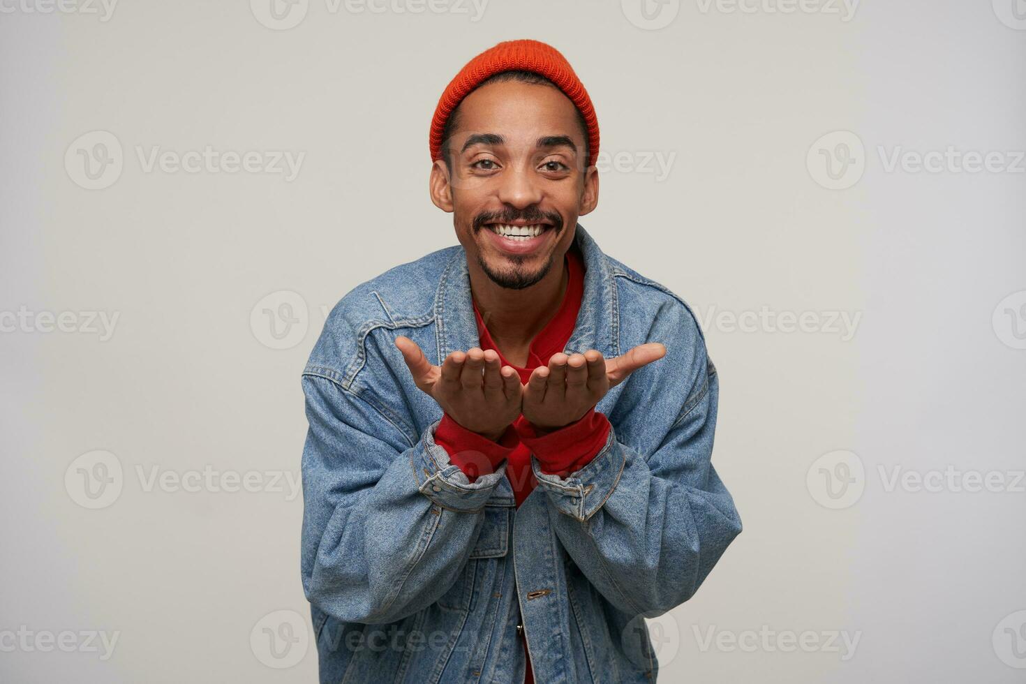 Happy young dark skinned bearded brunette male looking positively at camera with charming smile and keeping hands raised, posing over white background in casual clothes photo