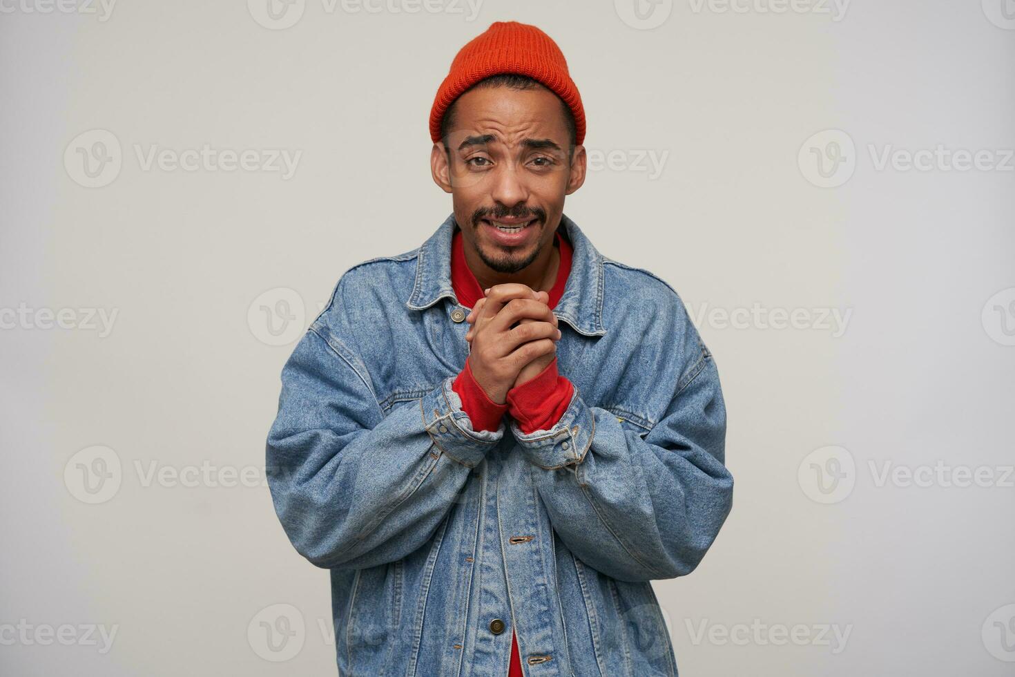 Indoor photo of young handsome bearded male with dark skin folding raised hands while standing over white background, wrinkling forehead while looking at camera