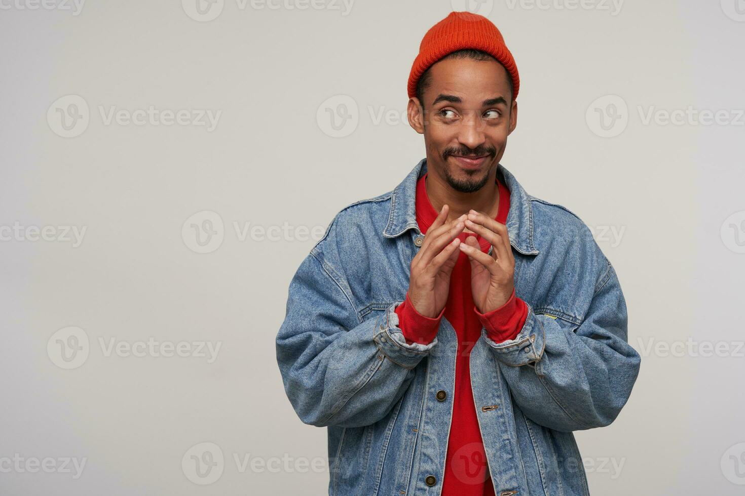Positive young brown-eyed bearded brunette guy with dark skin looking playfully aside with light smile and folding raised hands, standing against white background photo