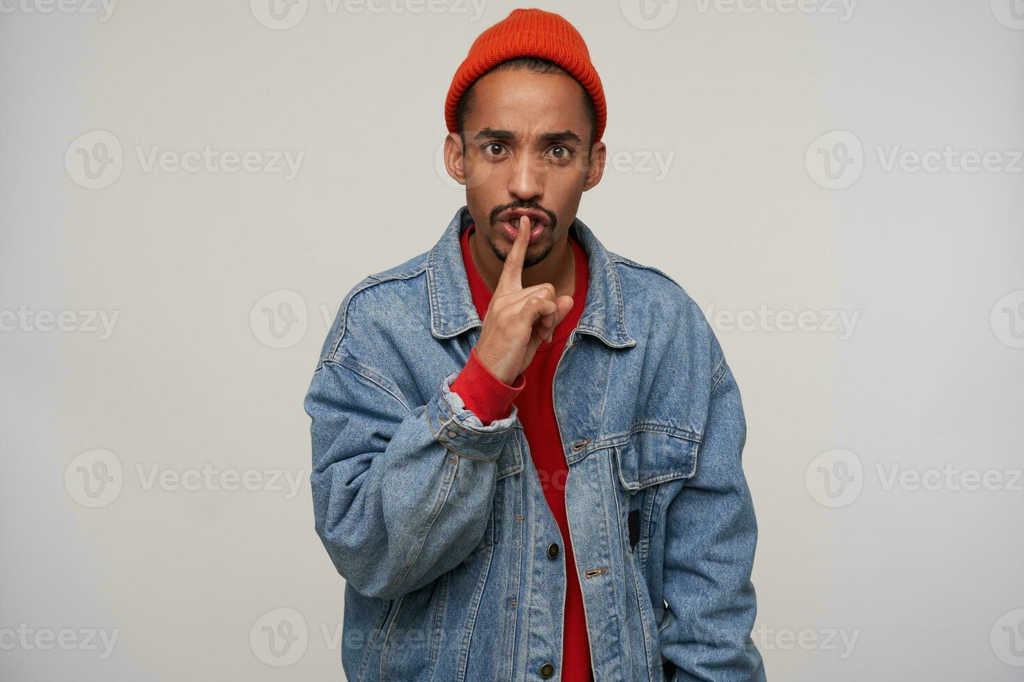 Serious young attractive dark skinned bearded male dressed in casual clothes holding forefinger on his mouth and frowning eyebrows while looking to camera, isolated over white background photo