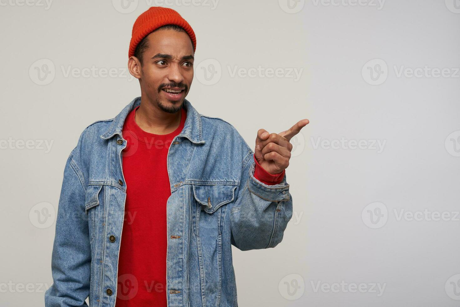 Photo of puzzled young dark skinned bearded guy frowning his eyebrows while looking aside with confused face, standing over white background with raised forefinger