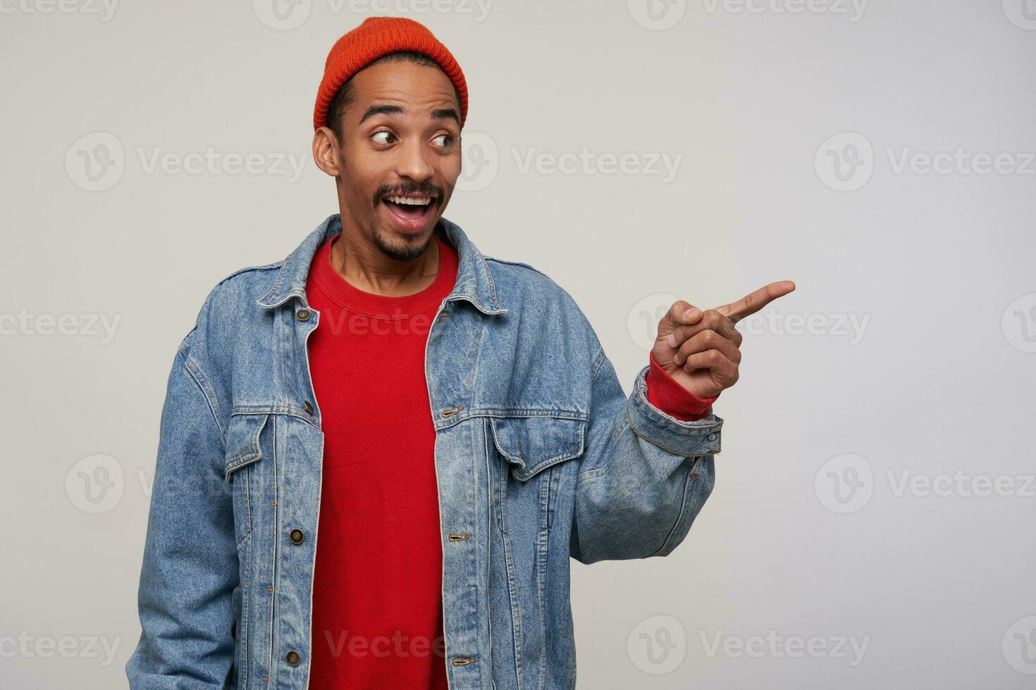 Cheerful young attractive bearded brown haired guy with dark skin showing aside with raised index finger and smiling joyfully while standing over white background photo