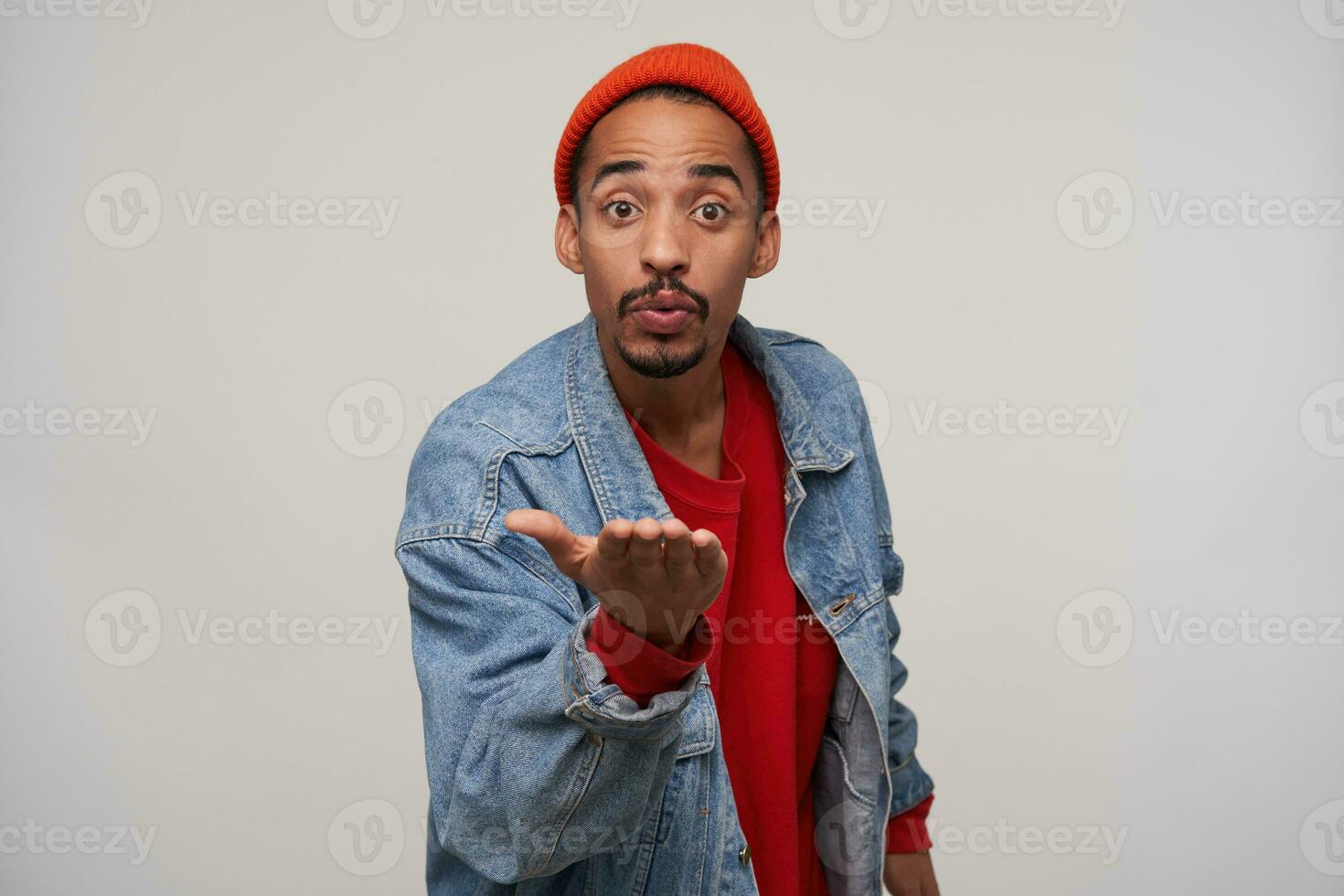 Positive young brown-eyed bearded brunette guy with dark skin holding raised palm and blowing air kiss to camera, dressed in red cap, pullover and blue jeans coat while posing over white background photo