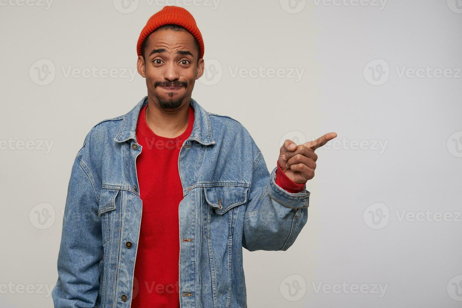 Studio photo of young dark skinned brunette man with beard getting air in his mouth and looking confusedly at camera, keeping index finger raised while posing over white background