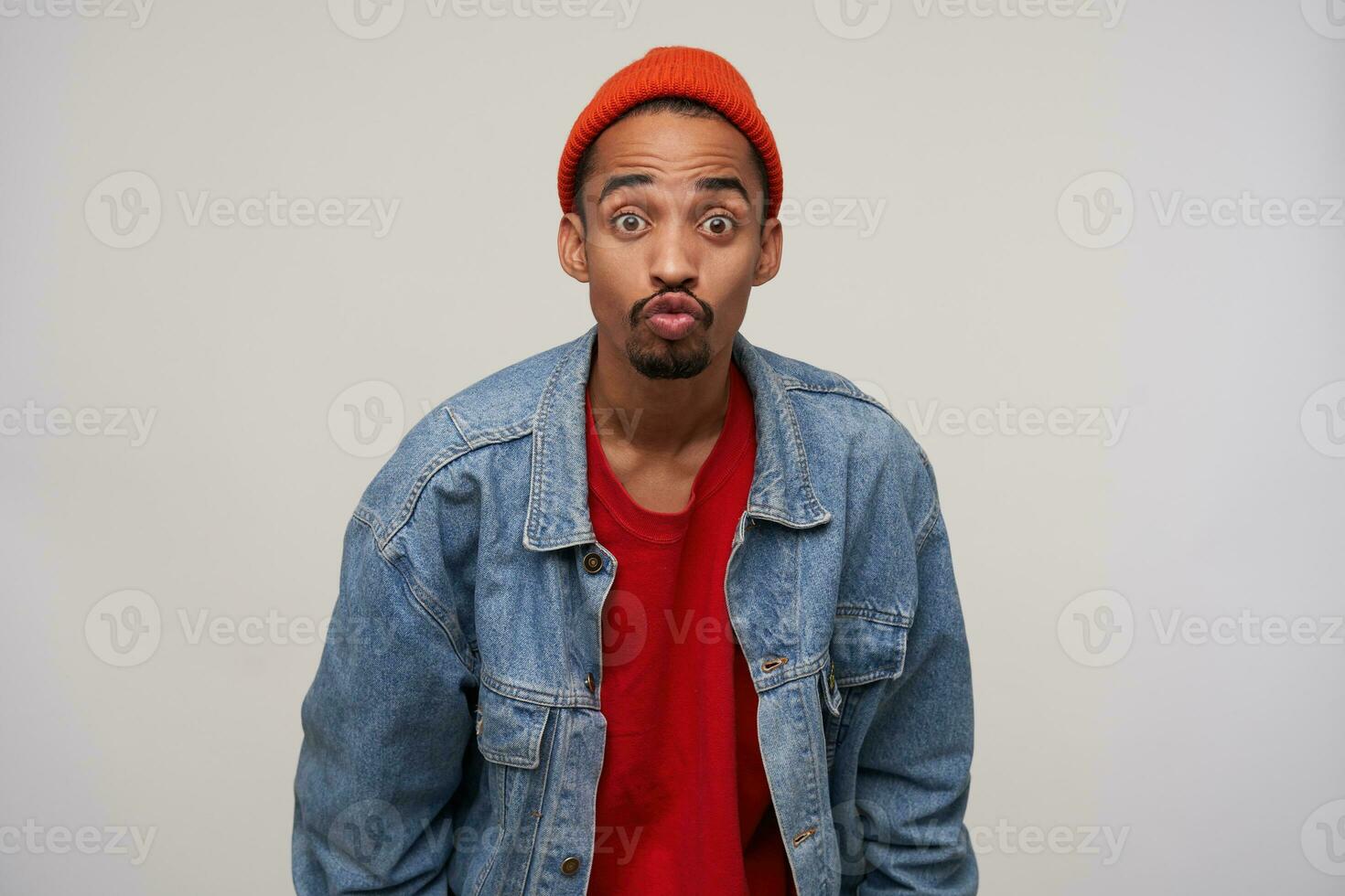 Funny shot of brown-eyed young handsome bearded brunette male with dark skin folding lips in air kiss while looking excitedly to camera, standing over white background photo
