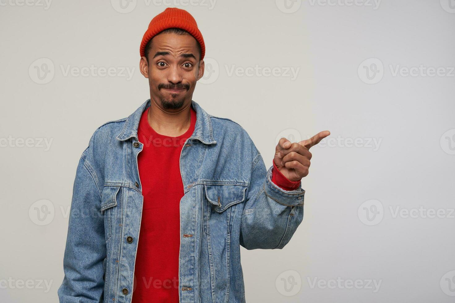 Confused young brown-eyed bearded dark haired male raising surprisedly eyebrows and puffing out cheeks while showing aside with index forefinger, isolated over white background photo