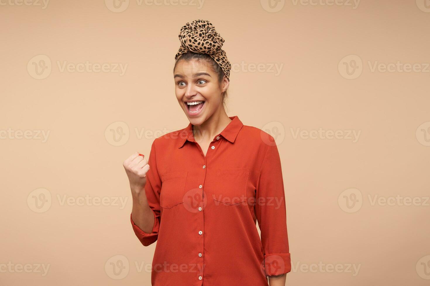 Overjoyed young attractive brunette female with natural makeup raising happily her hand while rejoicing about something, standing over beige background photo