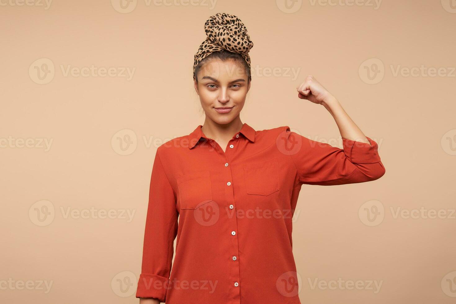 Positive young pretty brunette woman with natural makeup squinting her eyes while looking at camera and keeping hand raised, posing over beige background photo