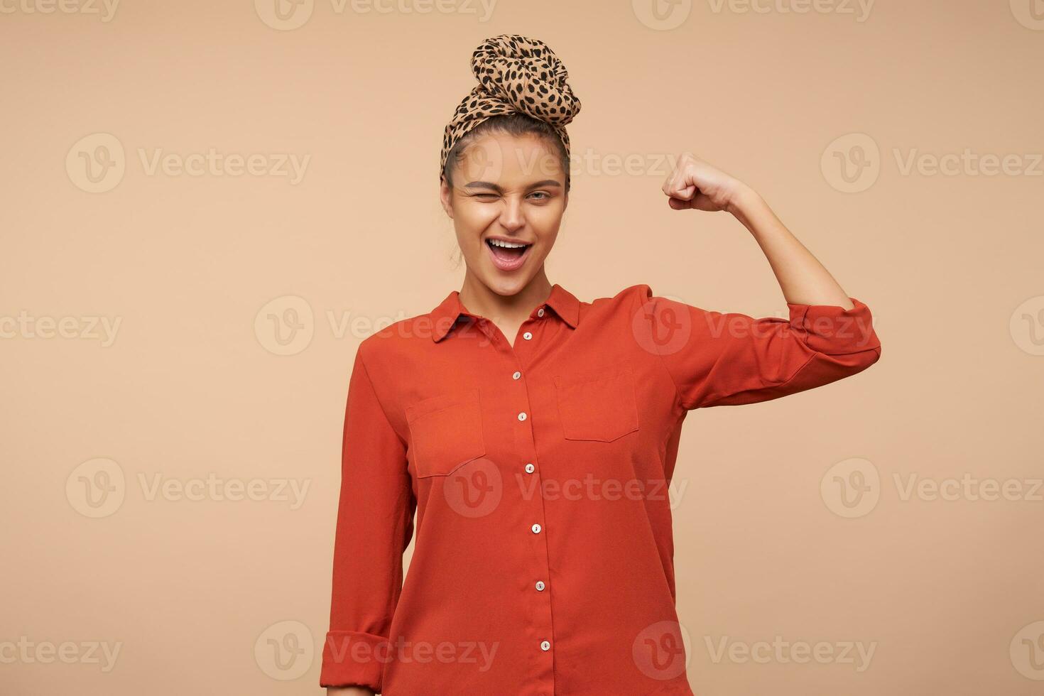 Cheerful young pretty brown haired lady with headband winking joyfully at camera while demonstrating her strong biceps with raised hand, isolated over beige background photo