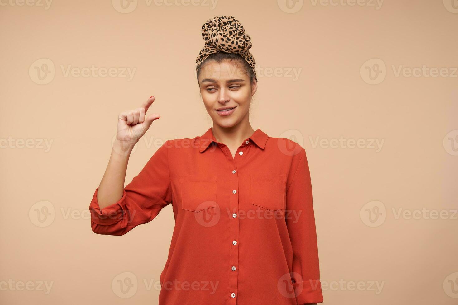 Puzzled young brown haired lady wearing headband in knot while standing over beige background, looking confusedly on her hand while showing small size with fingers photo
