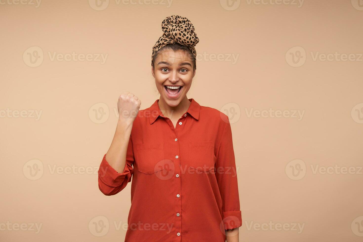 Excited young happy pretty brown haired woman with headband looking emotionally at camera while raising hand in yes gesture, isolated over beige background photo