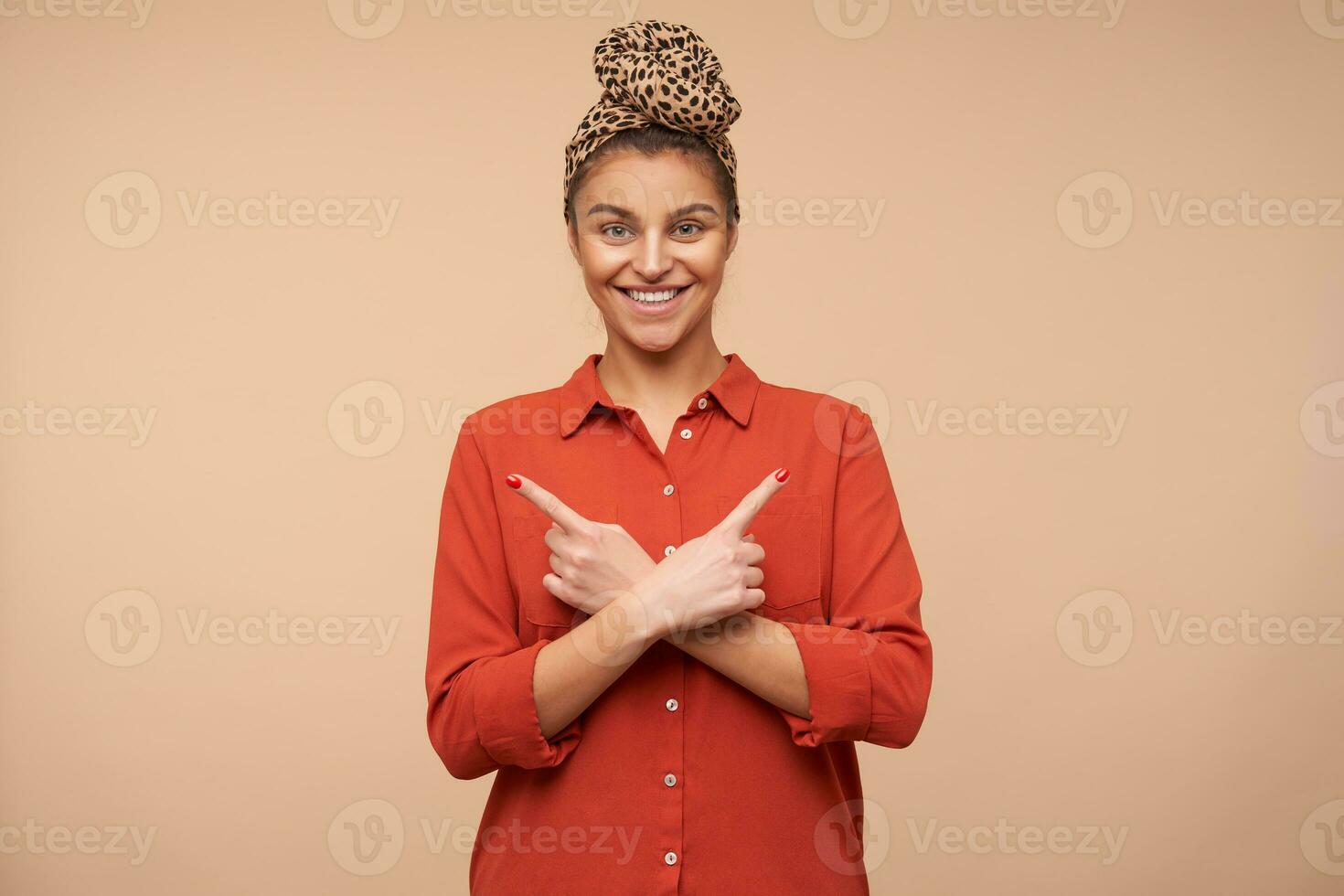 Happy young attractive brunette woman with headband smiling widely while pointing with forefingers in different directions, isolated over beige background photo