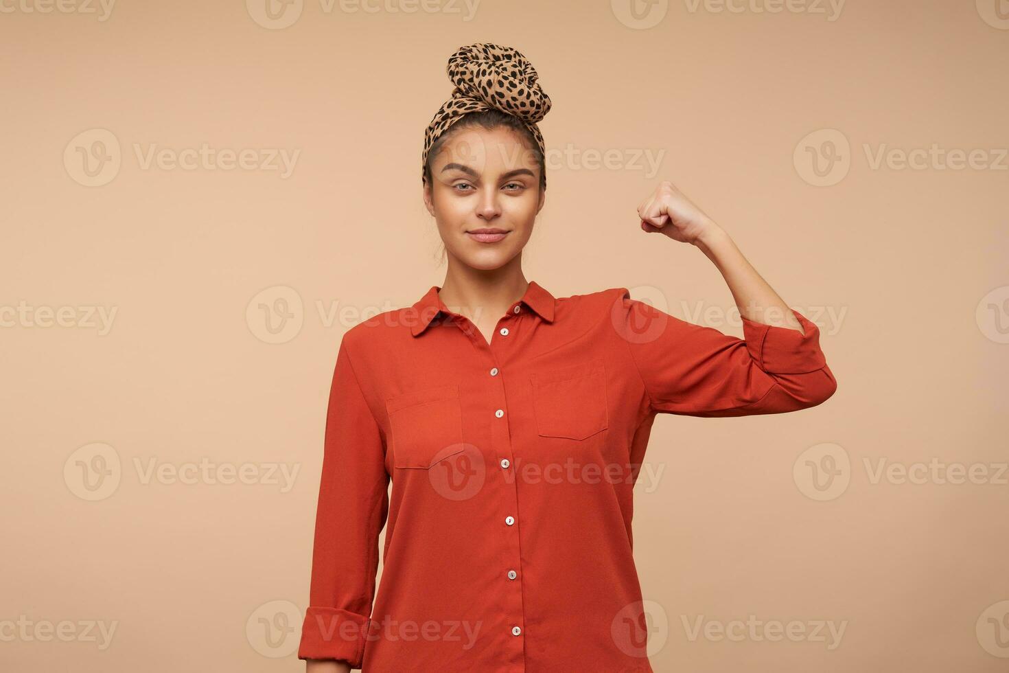Indoor photo of glad young brown haired woman dressed in red shirt raising her hand and looking attentively at camera with light smile, isolated over beige background
