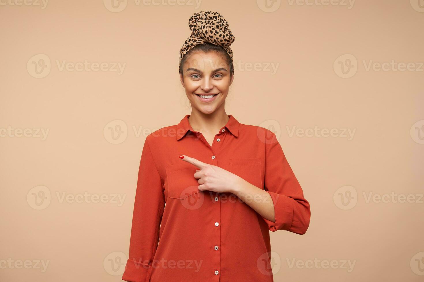 Cheerful young green-eyed brunette female smiling gladly while looking at camera and showing aside with index finger, isolated over beige background in casual wear photo