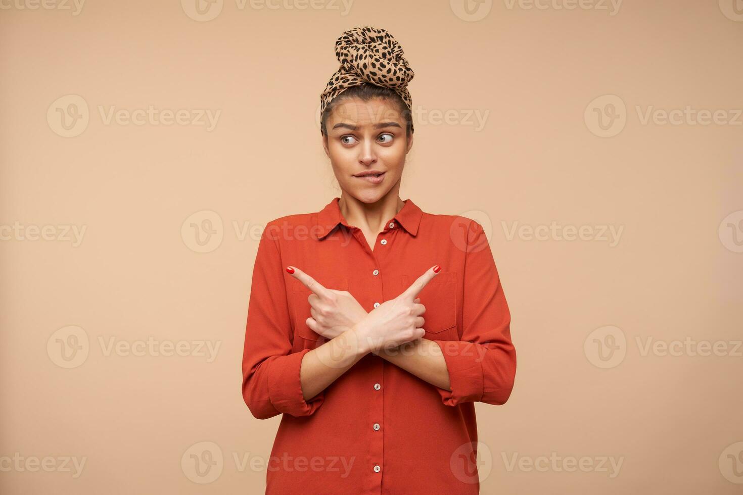 Bewildered young pretty brunette woman with headband biting worringly underlip while showing in different directions with index fingers, posing over beige background photo