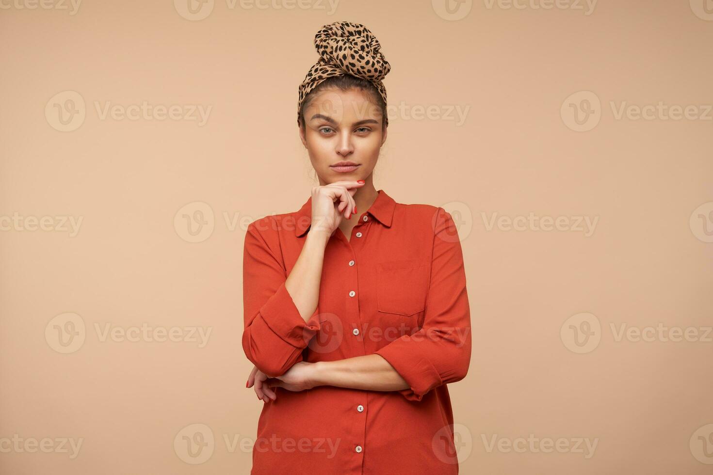 Pensive young pretty brunette lady squinting her eyes while looking thoughtfully at camera and holding chin with raised hand, isolated over beige background photo