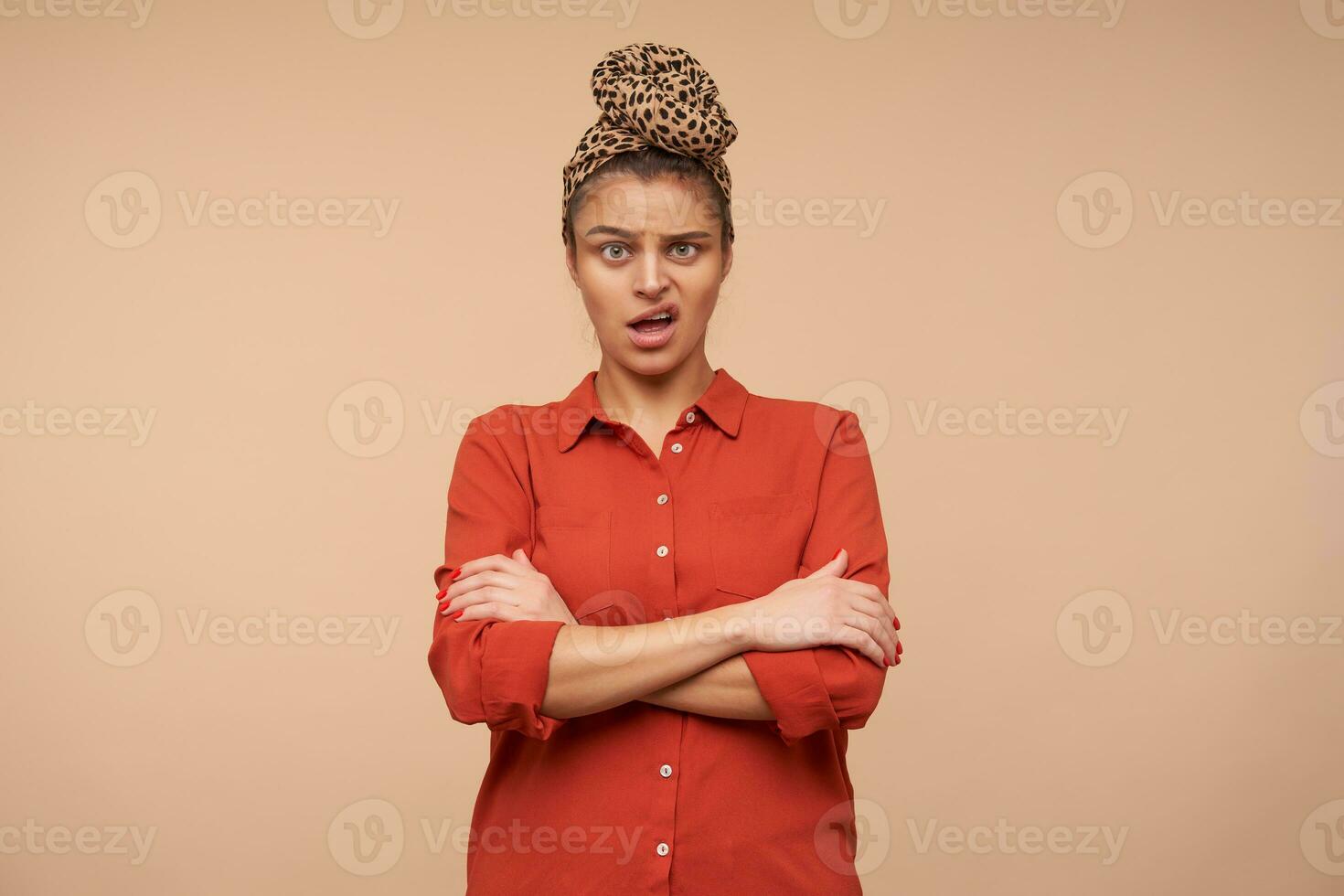 Studio photo of bewildered young brunette lady with headband grimacing confusedly her face while looking at camera and cossing hands on chest, isolated over beige background