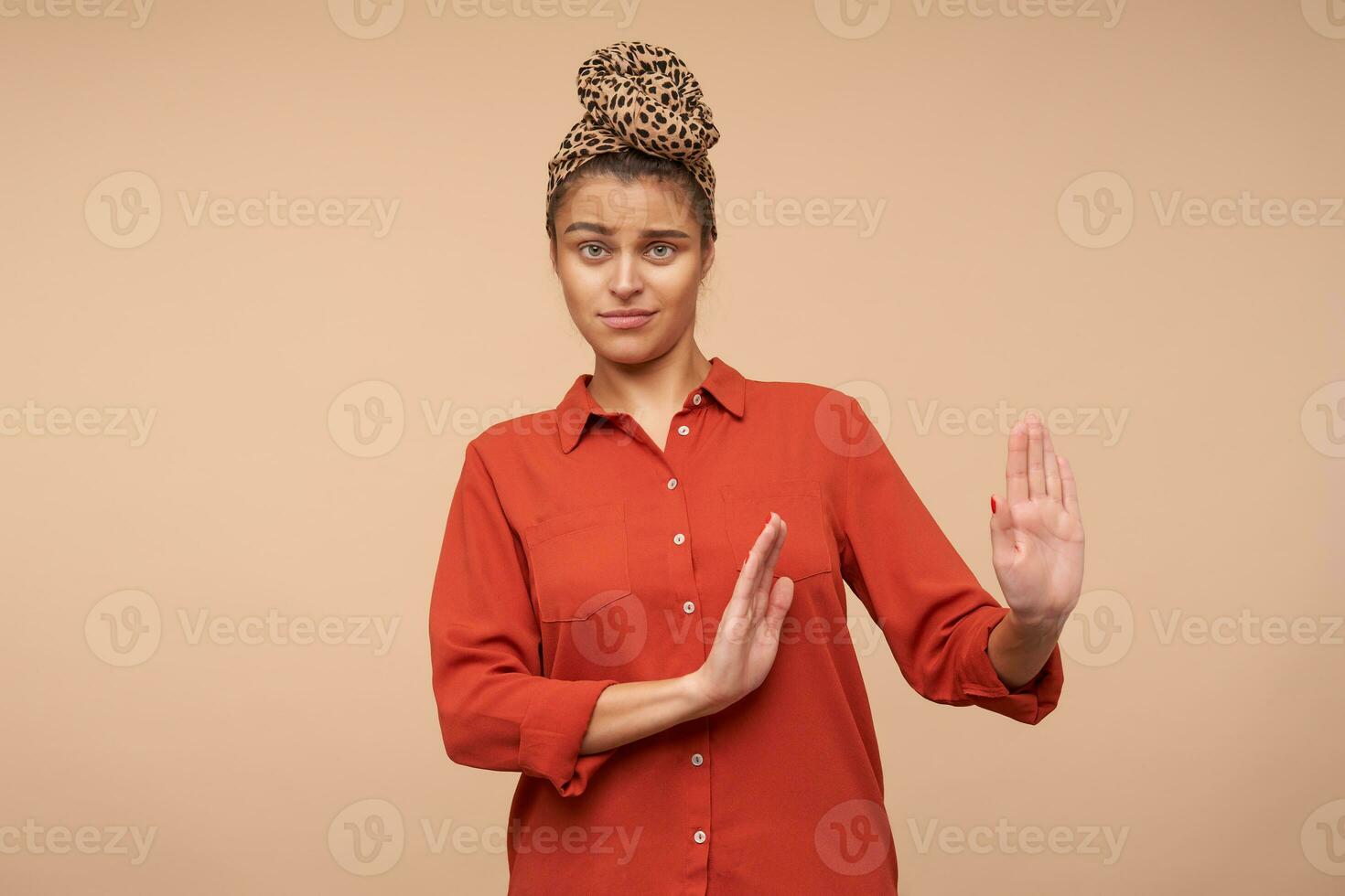 Indoor photo of young pretty green-eyed brunette lady keeping her lips folded while looking at camera and raising her palms in refusing gesture, isolated over beige background