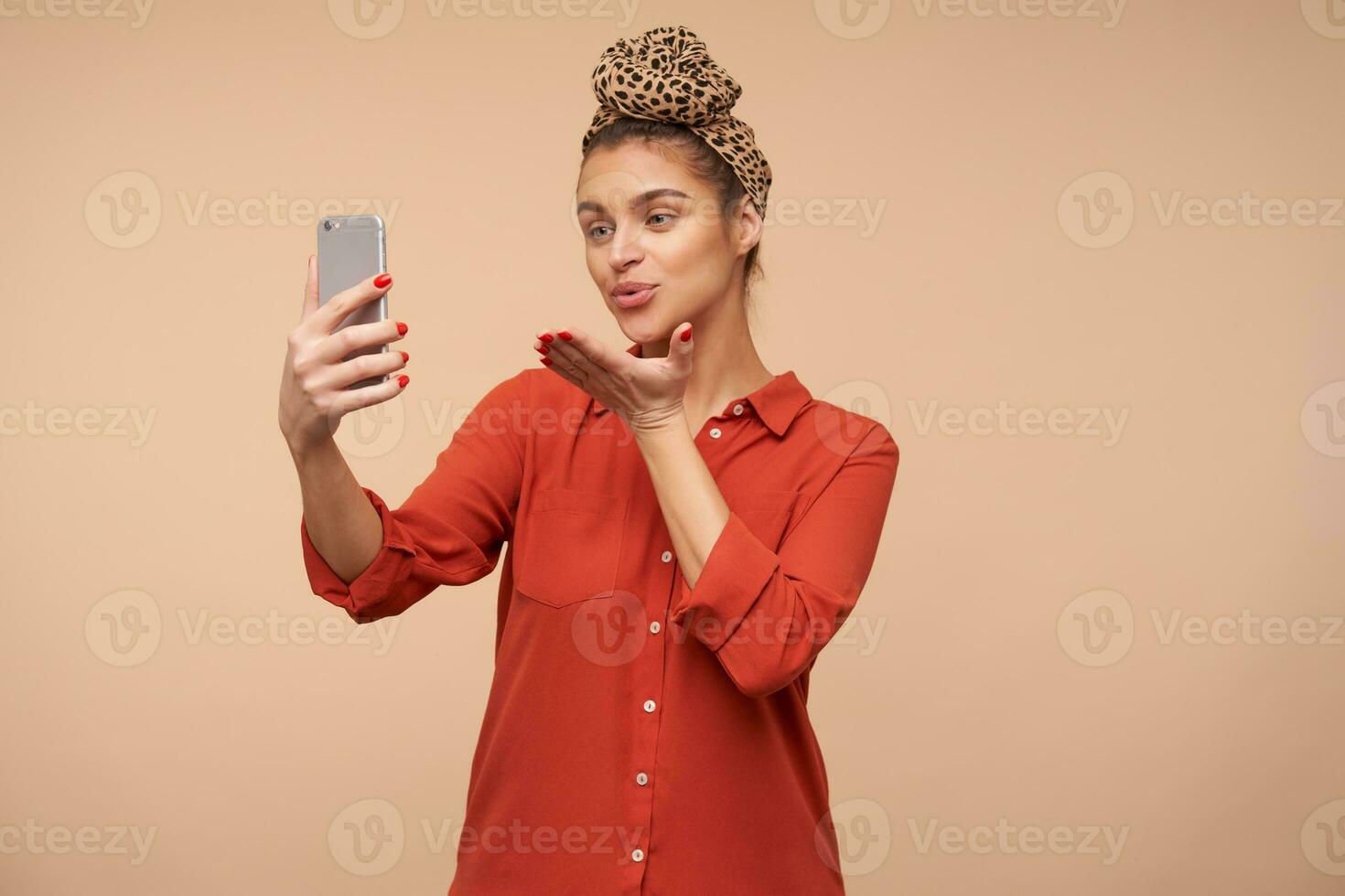 positivo joven atractivo morena hembra vestido en rojo camisa plegable su labios y acuerdo palma elevado mientras soplo aire Beso a cámara, aislado terminado beige antecedentes foto