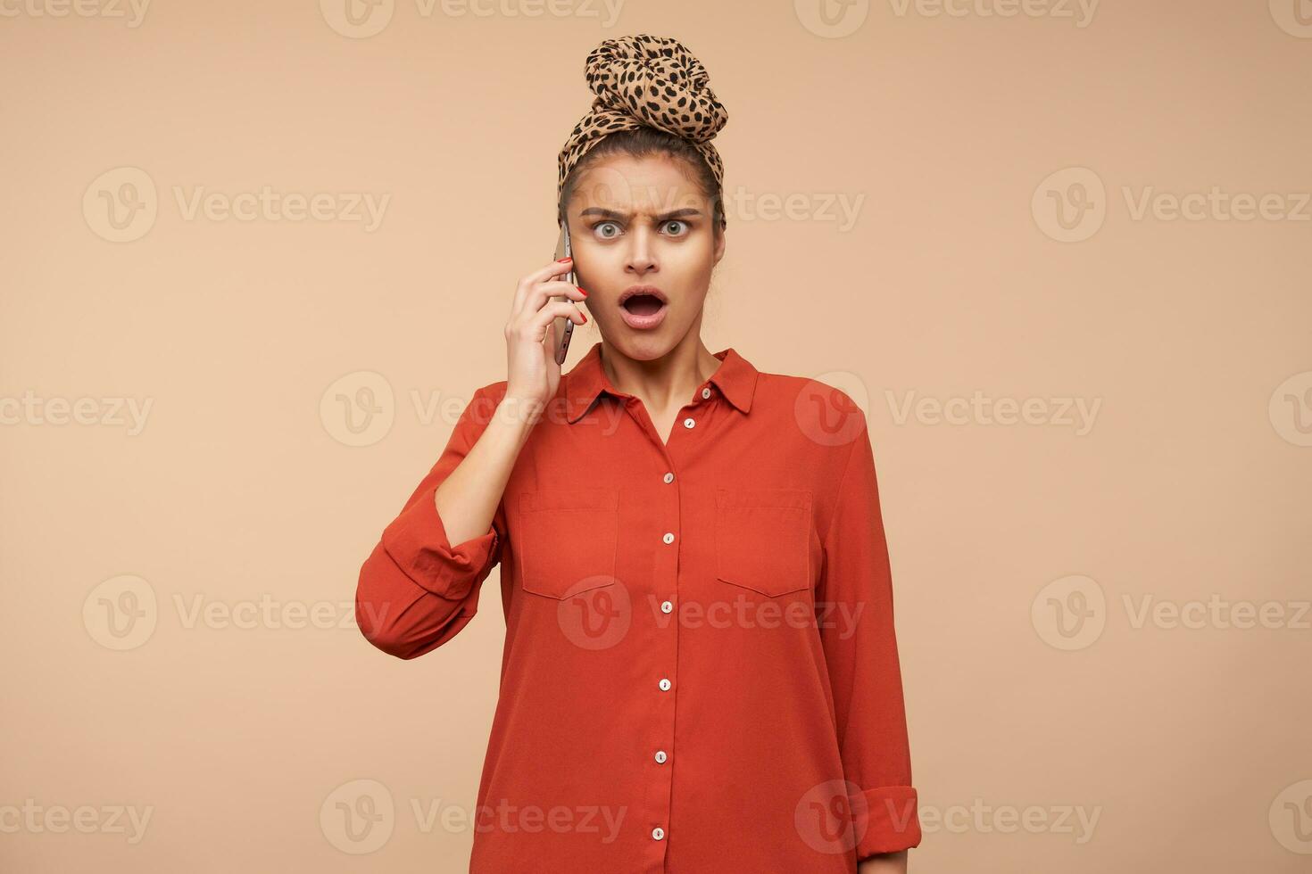 Indoor photo of young shocked brunette woman with natural makeup frowning her eyebrows while having tense phone conversation and looking amazedly at camera, isolated over beige background