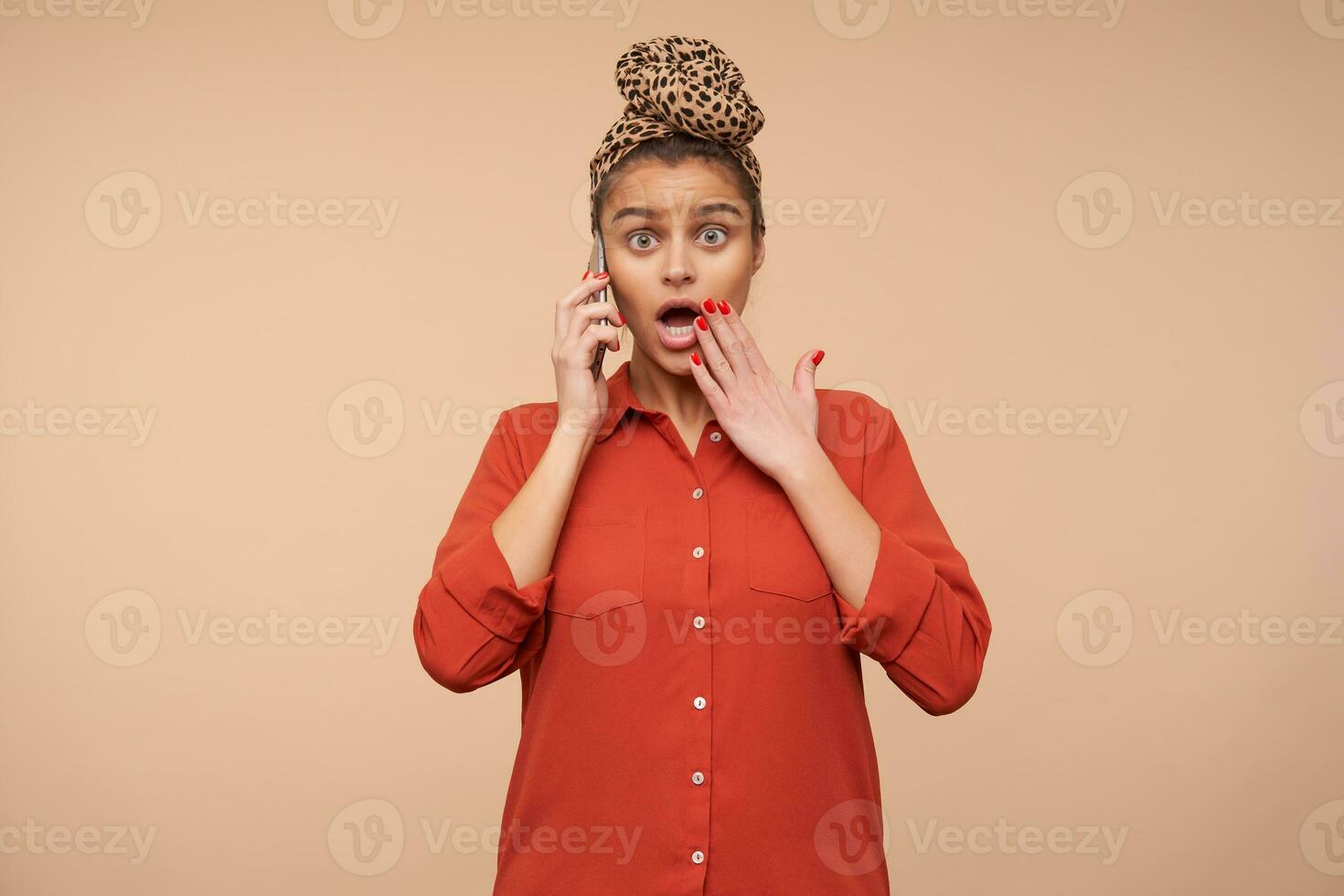 Bemused young attractive brunette woman with headband rounding dazedly her green eyes and raising emotionally hand to her mouth while hearing surpriseing news on phone photo