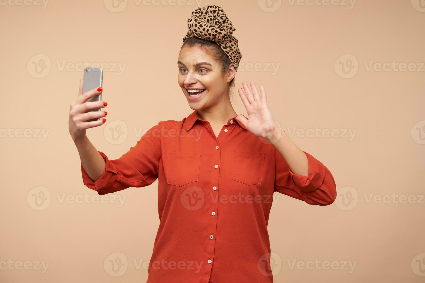 Happy young lovely brown haired female with headband smiling cheerfully at camera of her phone and raisinghand in hello gesture while having video call, posing over beige background photo