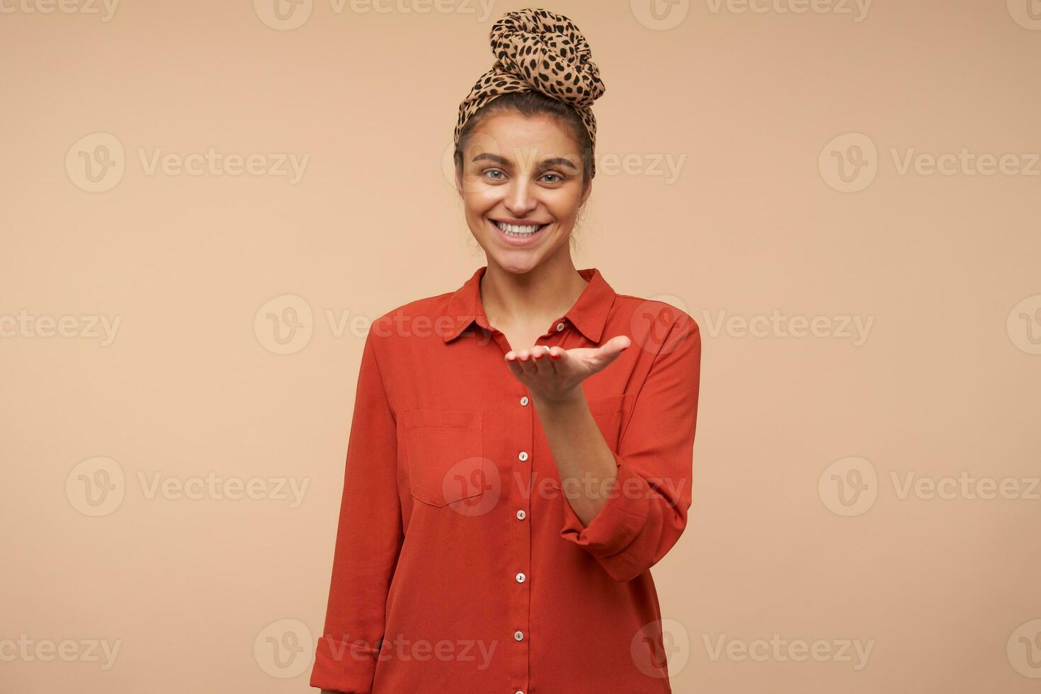 Happy young pretty brunette lady with natural makeup looking at camera with charming smile and raising up her palm, posing over beige background in red shirt photo