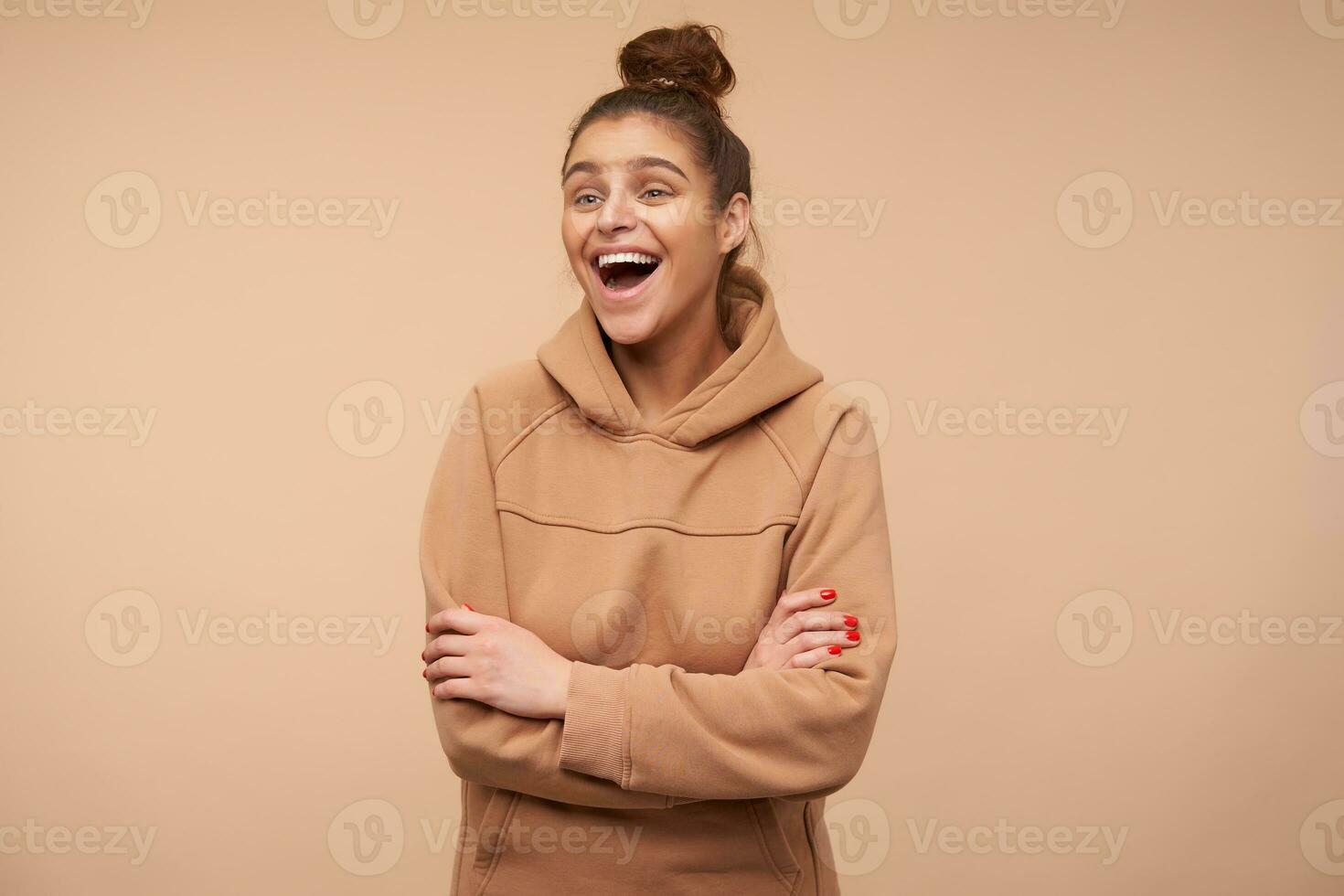 Indoor photo of young lovely brown haired lady with red manicure being in high spirit and laughing happily while standing over beige background with crossed arms