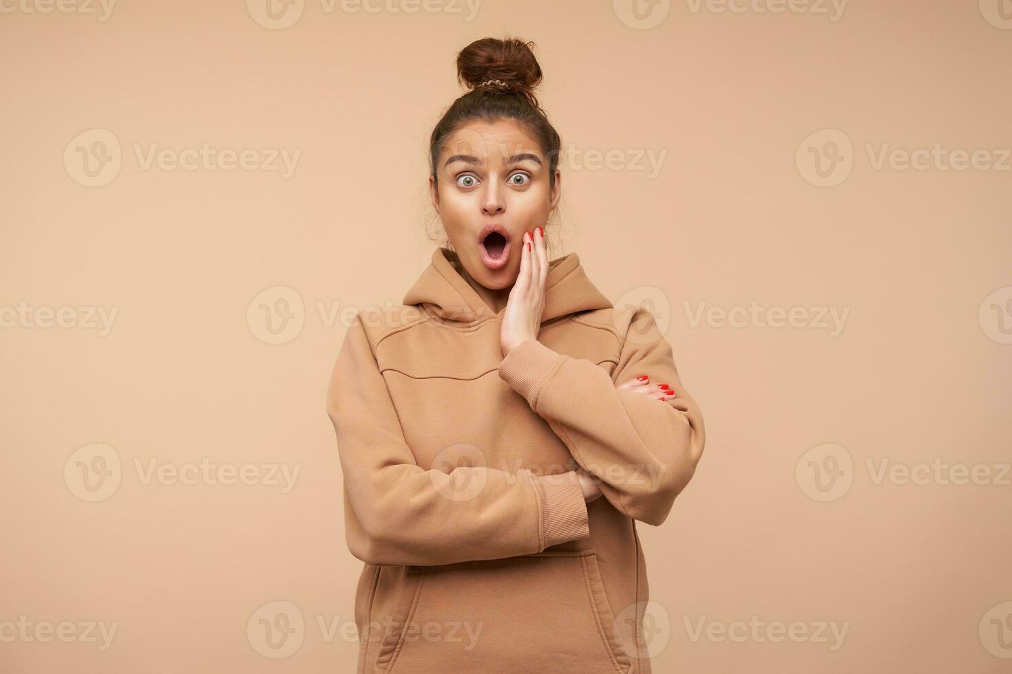 Dazed young green-eyed brunette female with bun hairstyle raising surprisedly her eyebrows while looking amazedly at camera and keeping palm on her cheek, posing over beige background photo
