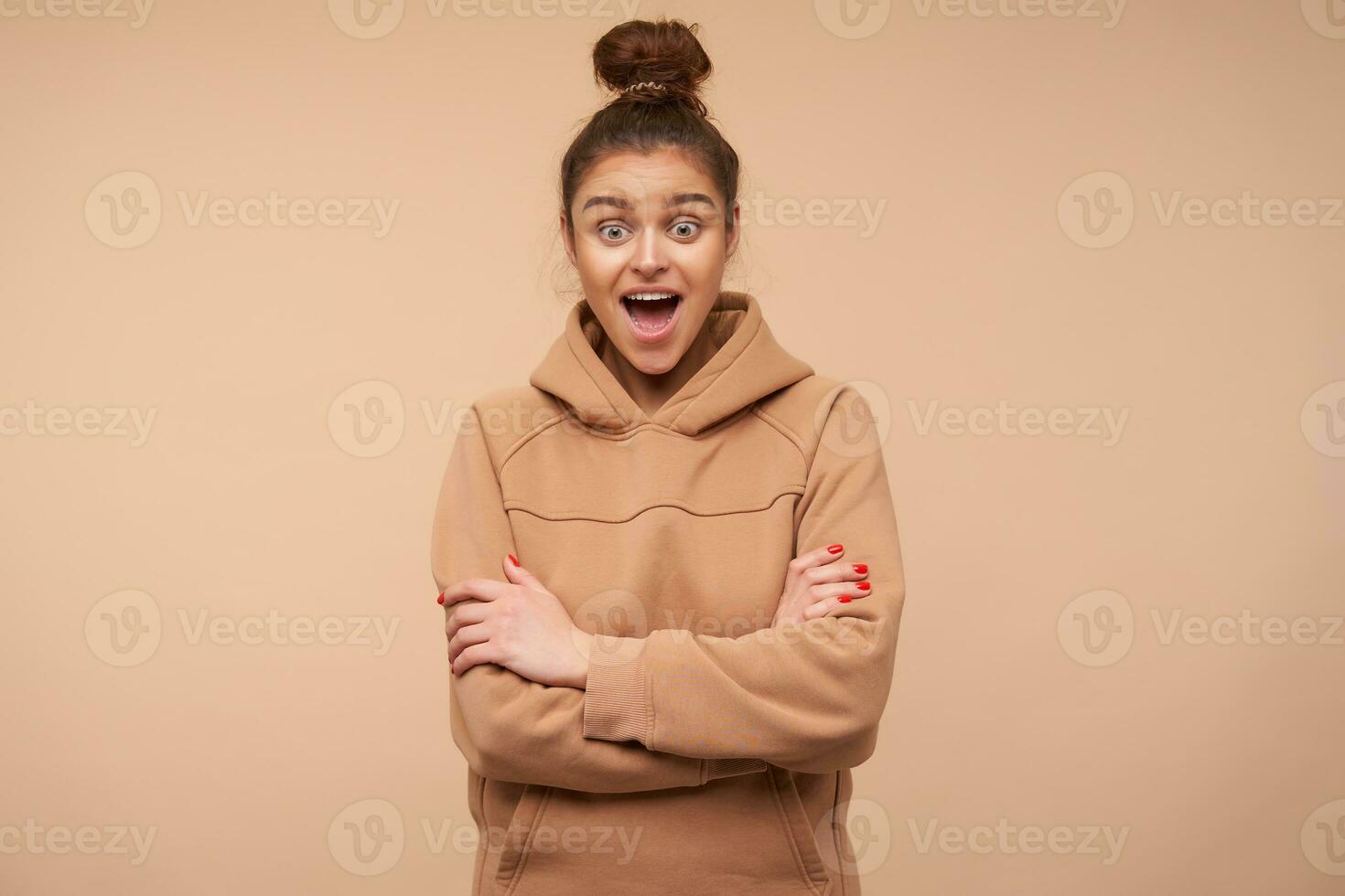 Bemused young green-eyed brunette woman dressed in nude sweatshirt looking excitedly ahead with wide mouth opened and folding hands on her chest, posing over beige background photo
