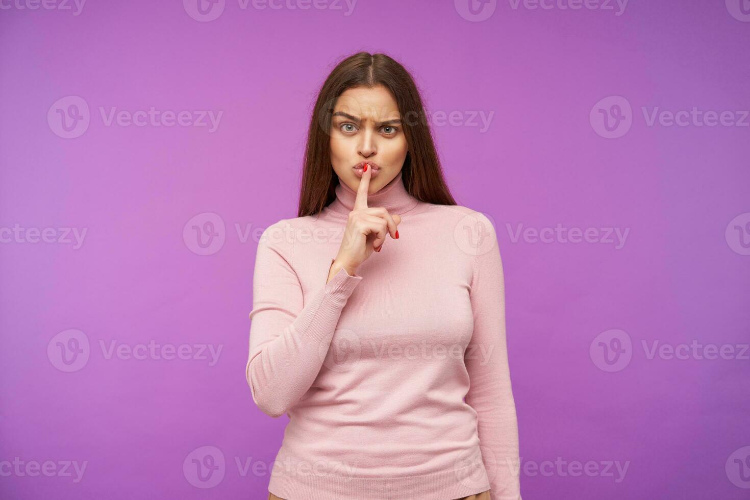 Serious young pretty brown haired woman with natural makeup frowning her eyebrows while looking severely at camera, raising hand with hush gesture while posing over purple background photo