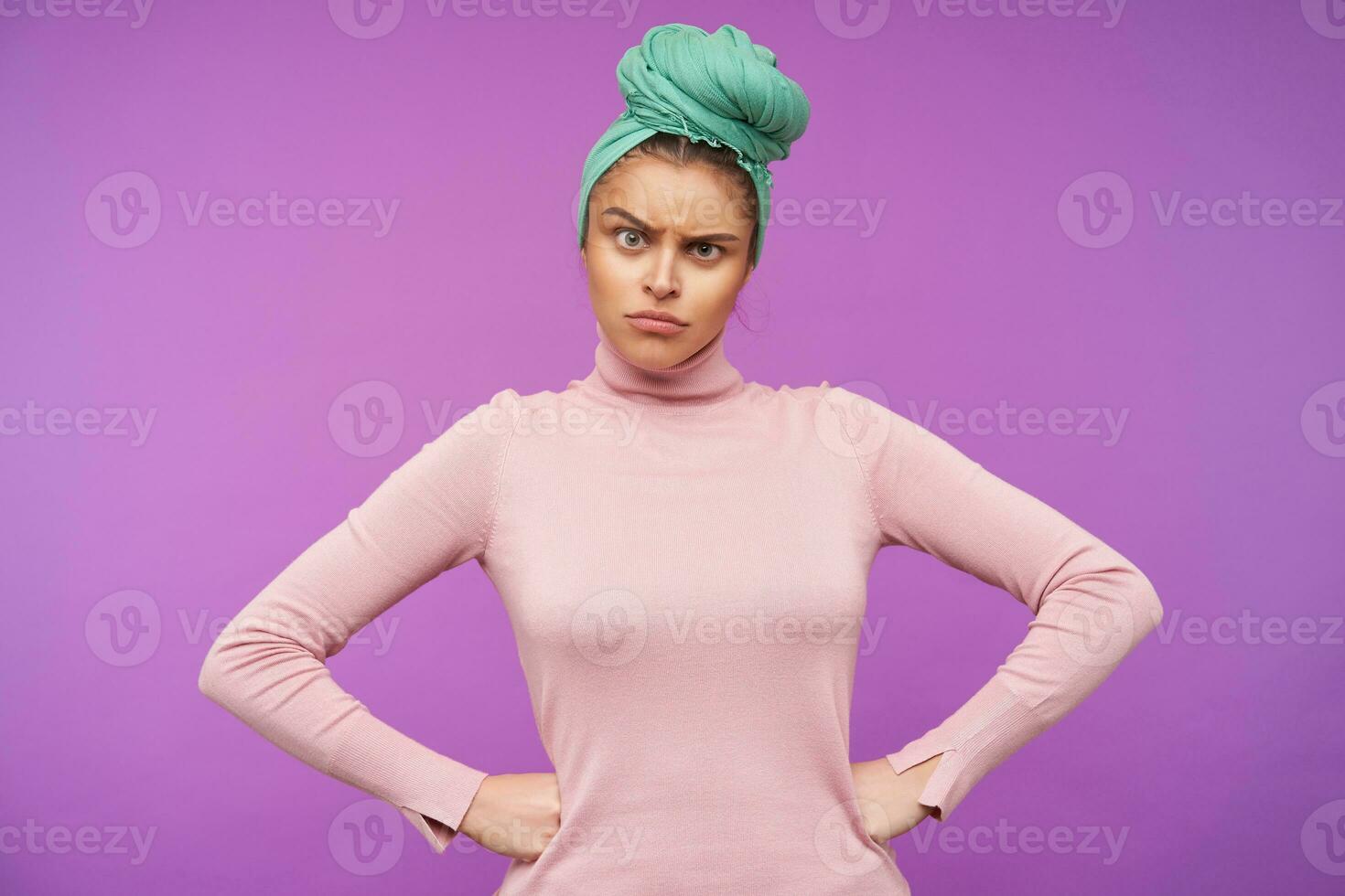 Studio photo of young serious brown haired woman with natural makeup frowning her eyebrows while looking crossly at camera, isolated over purple background