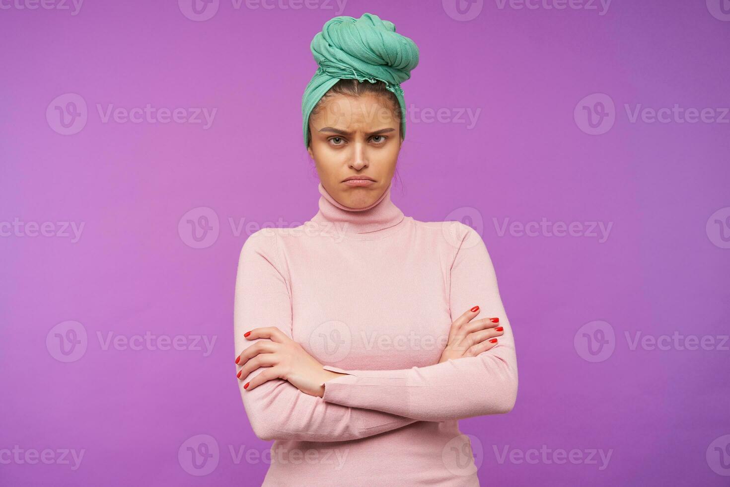 Offended young brown haired female with natural makeup folding hands on her chest while looking sadly at camera, posing over purple background in elegant wear photo